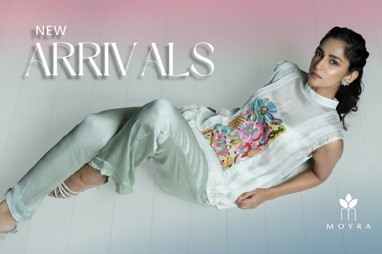 Woman wearing a colorful embroidered top and light green pants on a white background with 'New Arrivals' text and Moyra logo.